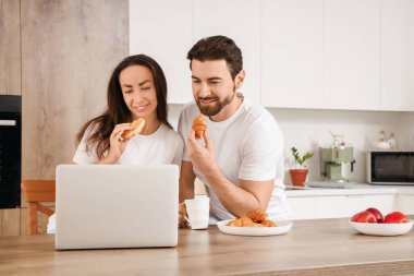 Mutfaktaki masada dizüstü bilgisayar kullanan kruvasan seven çift.
