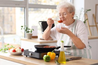 Kıdemli kadın mutfakta kızarmış yemek tadıyor.
