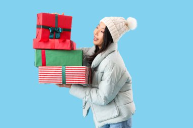 Mavi arka planda Noel hediyeleri olan kışlık giysili genç Asyalı kadın.