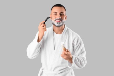 Gri arka planda yüzünü tıraş eden bornozlu genç bir adam.