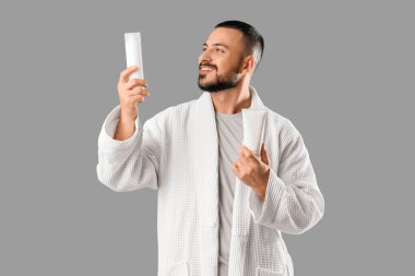 Gri arka planda cilt bakım kozmetiği olan bornozlu genç bir adam.