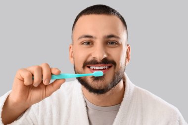 Gri arka planda dişlerini fırçalayan bornozlu genç adam.