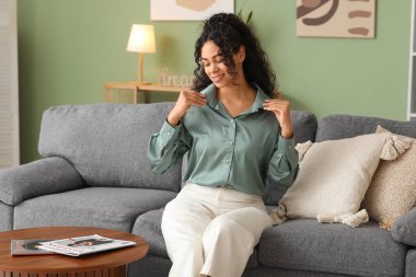 Evdeki kanepede oturan şık turkuaz gömlekli güzel Afro-Amerikalı kadın.