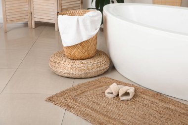 Yumuşak terlikler hasır halıda modern banyo küvetinin yanında.