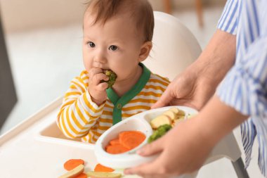 Anneciğiyle birlikte yüksek sandalyede brokoli yiyen tatlı bir bebek.