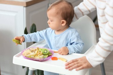 Yüksek sandalyede meyveleri olan sevimli küçük bebek ve evde annesi.