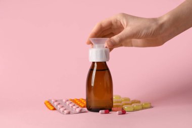 Female hand with bottle of cough syrup and different pills on pink background clipart