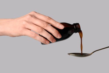 Woman pouring cough syrup from bottle into spoon on grey background clipart