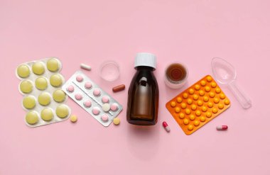 Bottle of cough syrup with measuring cups, spoon and different pills on pink background clipart