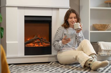 Elinde bir fincan kakao ve cep telefonu olan genç bir kadın şöminenin yanında oturuyor.