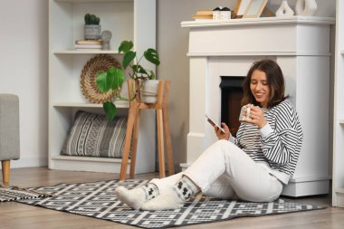 Elinde bir fincan kakao ve cep telefonu olan genç bir kadın şöminenin yanında oturuyor.