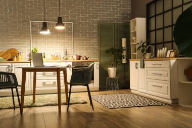 Karanlık mutfağın içinde tezgahlar, yemek masası ve akşamları parlayan lambalar.