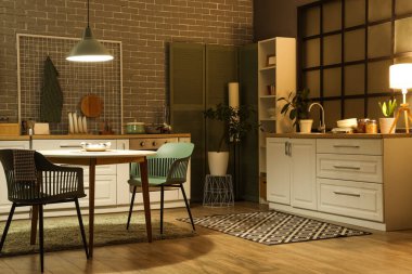 Karanlık mutfağın içinde tezgahlar, yemek masası ve akşamları parlayan lambalar.