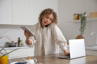 Bilgisayarı ve tablet bilgisayarı olan stresli genç bir kadın mutfakta cep telefonuyla konuşuyor.
