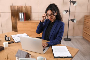 Beautiful African-American lawyer working with laptop at table in office clipart