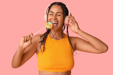 Lolipopu olan genç Afro-Amerikalı kadın pembe arka planda müzik dinliyor.