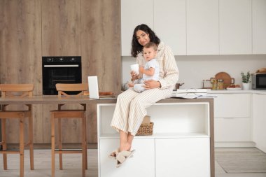 Kahve fincanı ile çalışan anne ve küçük bebeği mutfaktaki masada oturuyor.