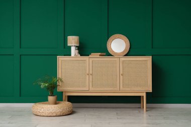 Yeşil duvarın yanında kitap, ayna ve masa lambası olan ahşap bir dolap.