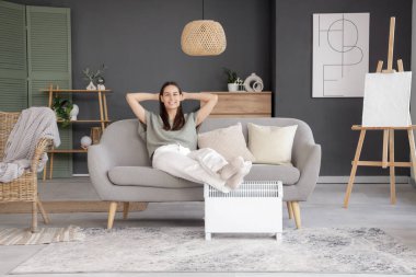 Oturma odasındaki elektrikli ısıtıcının yanındaki kanepede oturan genç kadın.