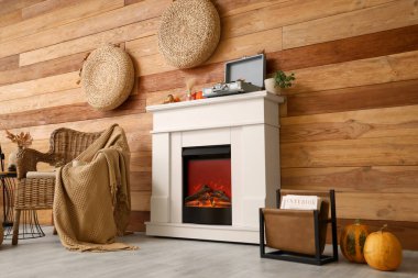 Hasır koltuğu, pikabı ve kahverengi ahşap duvarın yanındaki modern şöminede sonbahar süslemeleri olan şık bir oda.