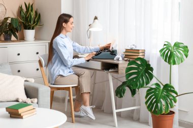 Daktilo, kitap ve buruşuk kağıtlarla dolu mutlu bir kadın yazar masada oturuyor.