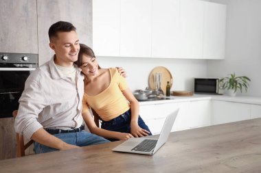 Genç çift mutfaktaki masada dizüstü bilgisayar kullanıyor.