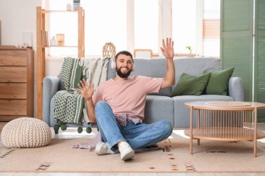 Kulaklıklı genç sakallı adam evdeki halıda oturuyor.