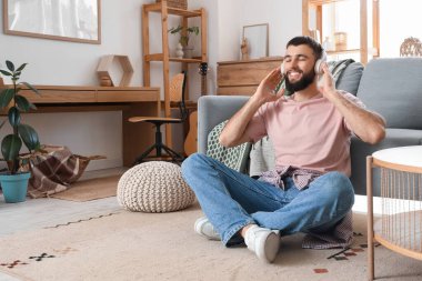 Kulaklıklı genç sakallı adam evdeki halıda oturuyor.