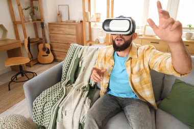 Genç sakallı adam evdeki kanepede VR gözlüğü kullanıyor.