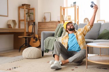 Fotoğraf makineli genç sakallı adam evde halının üstünde selfie çekiyor.