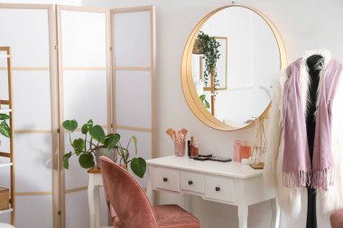 Kozmetik ürünleri, şık giysiler, katlanan ekran ve odada asılı ayna olan masa.