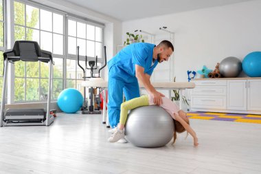 Erkek fizyoterapist rehabilitasyon merkezinde fitball oynayan küçük bir kızla çalışıyor.