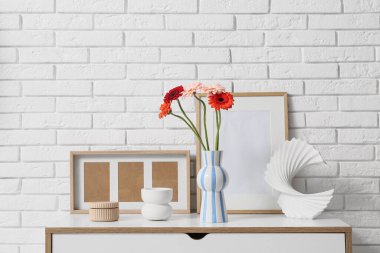 İçinde Gerbera çiçekleri olan vazo, fotoğraf çerçevesi ve çekmecelerde beyaz tuğla duvarın yanında duran heykel.