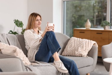 Evdeki kanepede bir fincan kahve içen genç bir kadın.