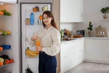 Genç bir kadın mutfaktaki buzdolabının yanında meyve suyu şişesi açıyor.