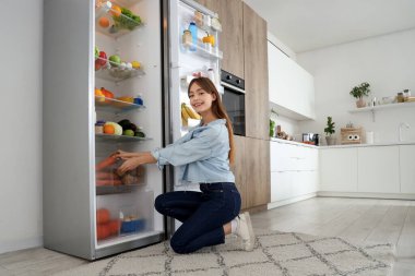 Genç bir kadın mutfaktaki açık buzdolabından havuç alıyor.