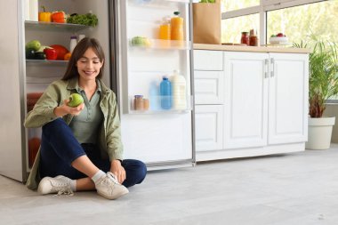 Elmalı genç bir kadın mutfakta açık buzdolabının yanında oturuyor.