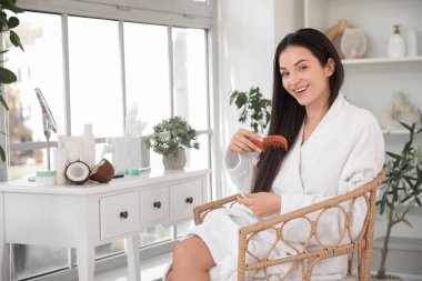 Kozmetik ürünleri ve Hindistan ceviziyle güzel, genç bir kadın banyoda saçını tarıyor.