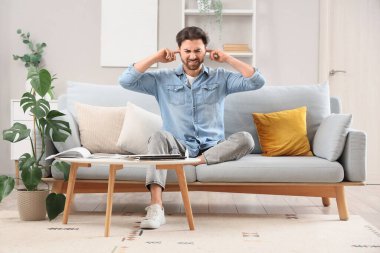 Evdeki kanepede kulaklarını kapatmakta zorlanan genç bir adam.