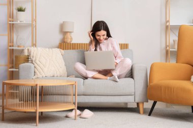 Çizgili pijamalı genç bir kadın evdeki kanepede dizüstü bilgisayarla çalışıyor.