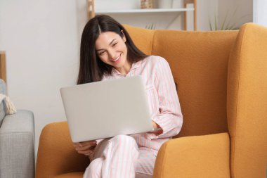 Çizgili pijamalı genç bir kadın evdeki koltukta dizüstü bilgisayarla çalışıyor.