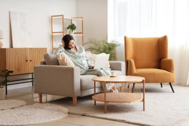 Yeşil pijamalı genç bir kadın evdeki kanepede dizüstü bilgisayarla çalışıyor.