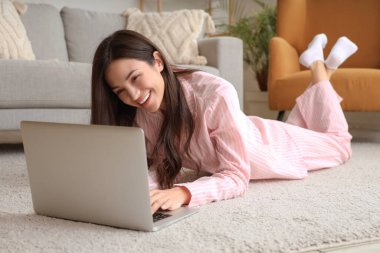 Pembe pijamalı kadın evde dizüstü bilgisayarla halıda çalışıyor.