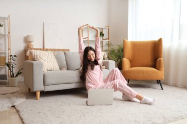 Evde dizüstü bilgisayarı halıya yayılmış pembe pijamalı serbest çalışan bir kadın.