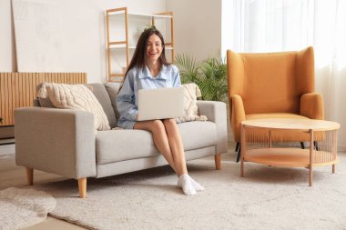 Evdeki kanepede dizüstü bilgisayarla çalışan mavi pijamalı genç bir kadın.