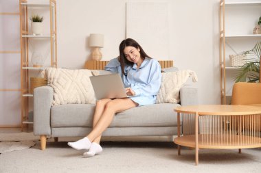 Evdeki kanepede dizüstü bilgisayarla çalışan mavi pijamalı genç bir kadın.