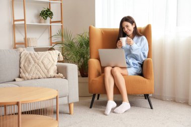 Kahve ve dizüstü bilgisayarlı mavi pijamalı genç bir kadın evde koltukta çalışıyor.