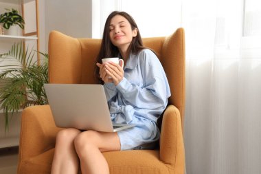 Kahve ve dizüstü bilgisayarlı mavi pijamalı genç bir kadın evde koltukta çalışıyor.