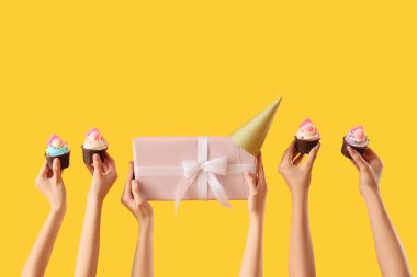 Doğum günü kekleri, parti şapkası ve sarı arka planda hediye kutusu olan bir sürü kadın eli.
