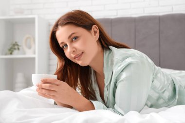 Genç ve güzel bir kadın bir fincan kahveyle yatakta yatıyor.